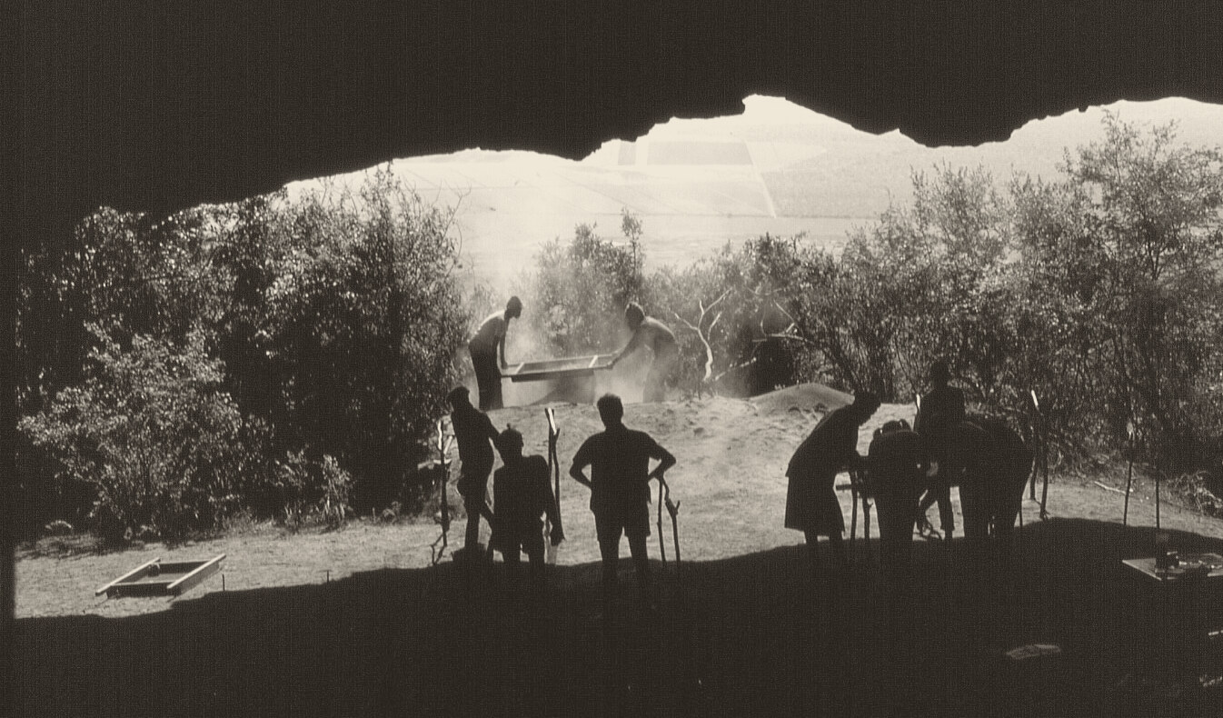 Black-and-white photograph from inside Border Cave looking out: the dark rock overhang frames a bright valley, and people stand in silhouette on the cave floor while two of them shake a sieve.