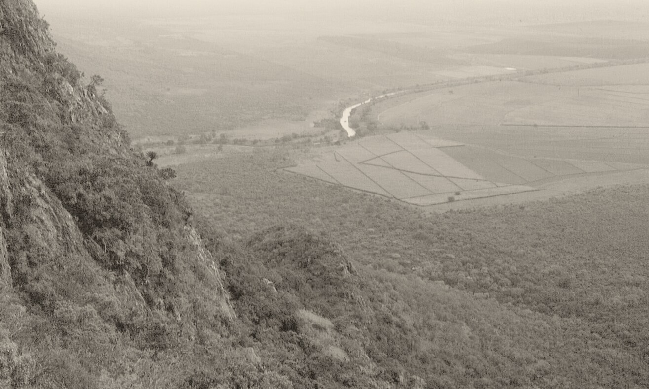 Black-and-white photograph from high on a rocky, bush-covered slope, looking down over a wide valley with a river, cleared fields and bushland stretching to the horizon.