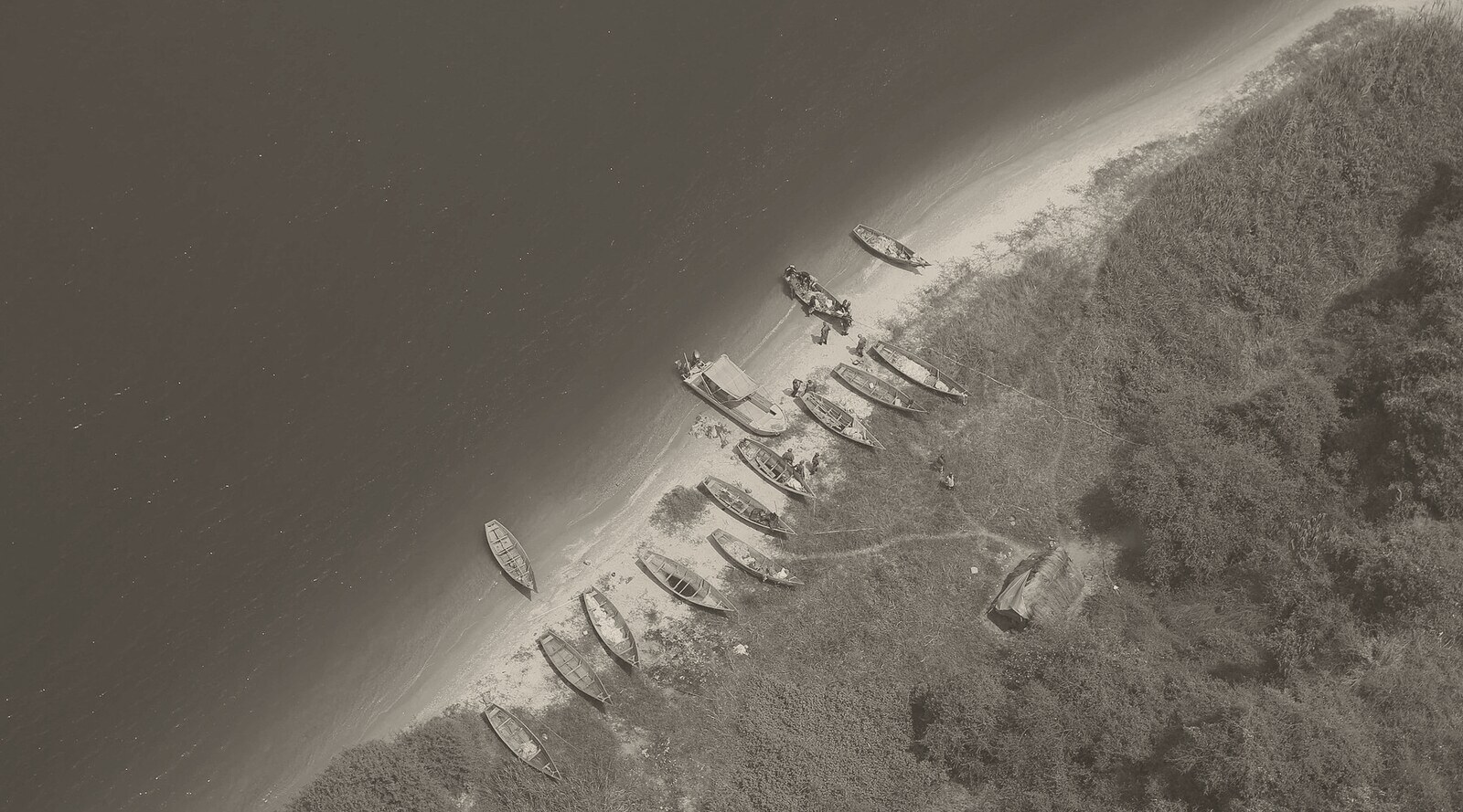 Aerial black-and-white photograph of a narrow sandy beach running diagonally between dark water and dense bush, with about fifteen wooden fishing boats pulled up on the sand, a few people beside them and a small thatched shelter behind.