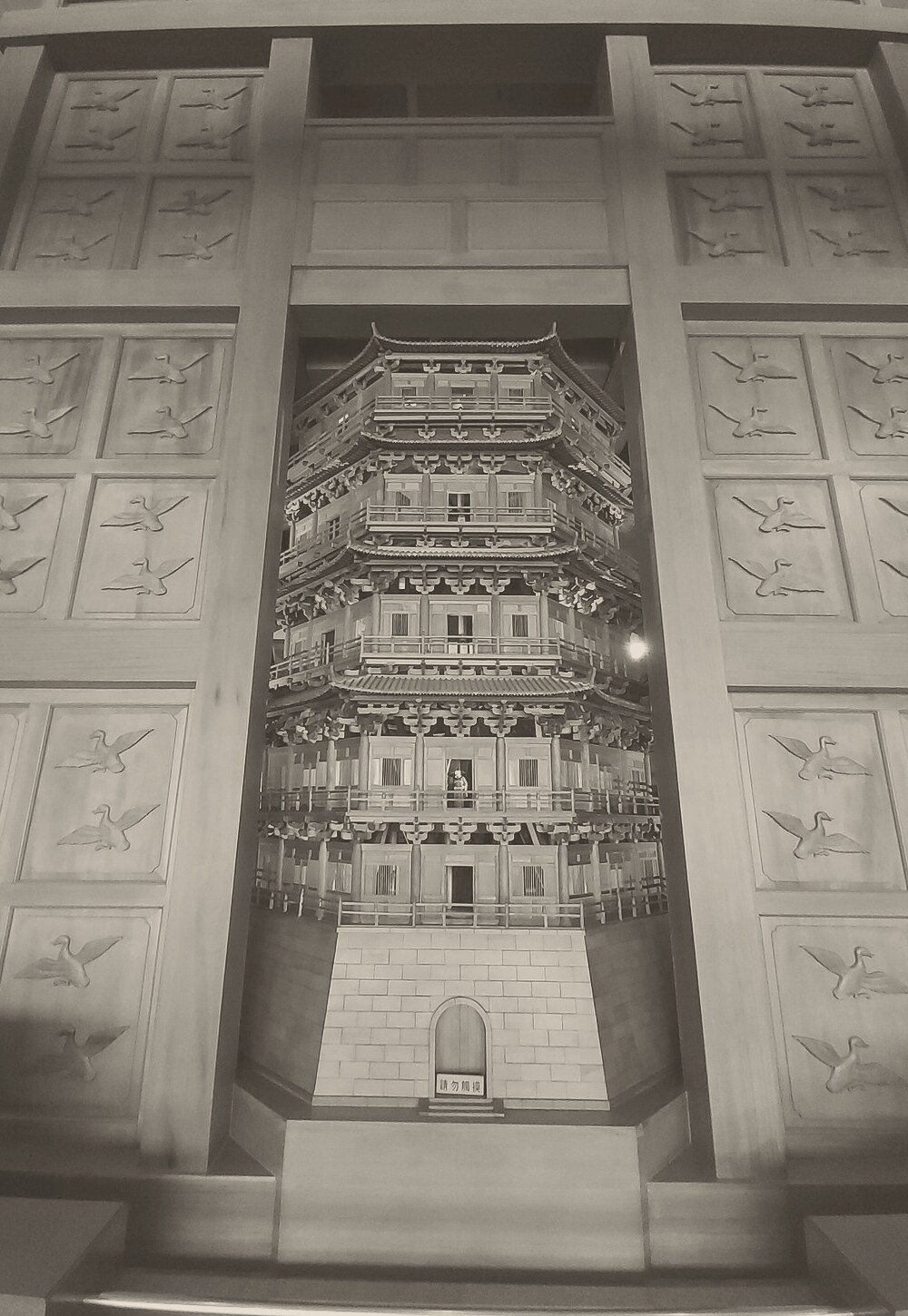 The front of a tall wooden tower in a museum: carved panels of birds frame a recess holding a five-storey pagoda on a stone base, with a small figure standing in one of its doorways.