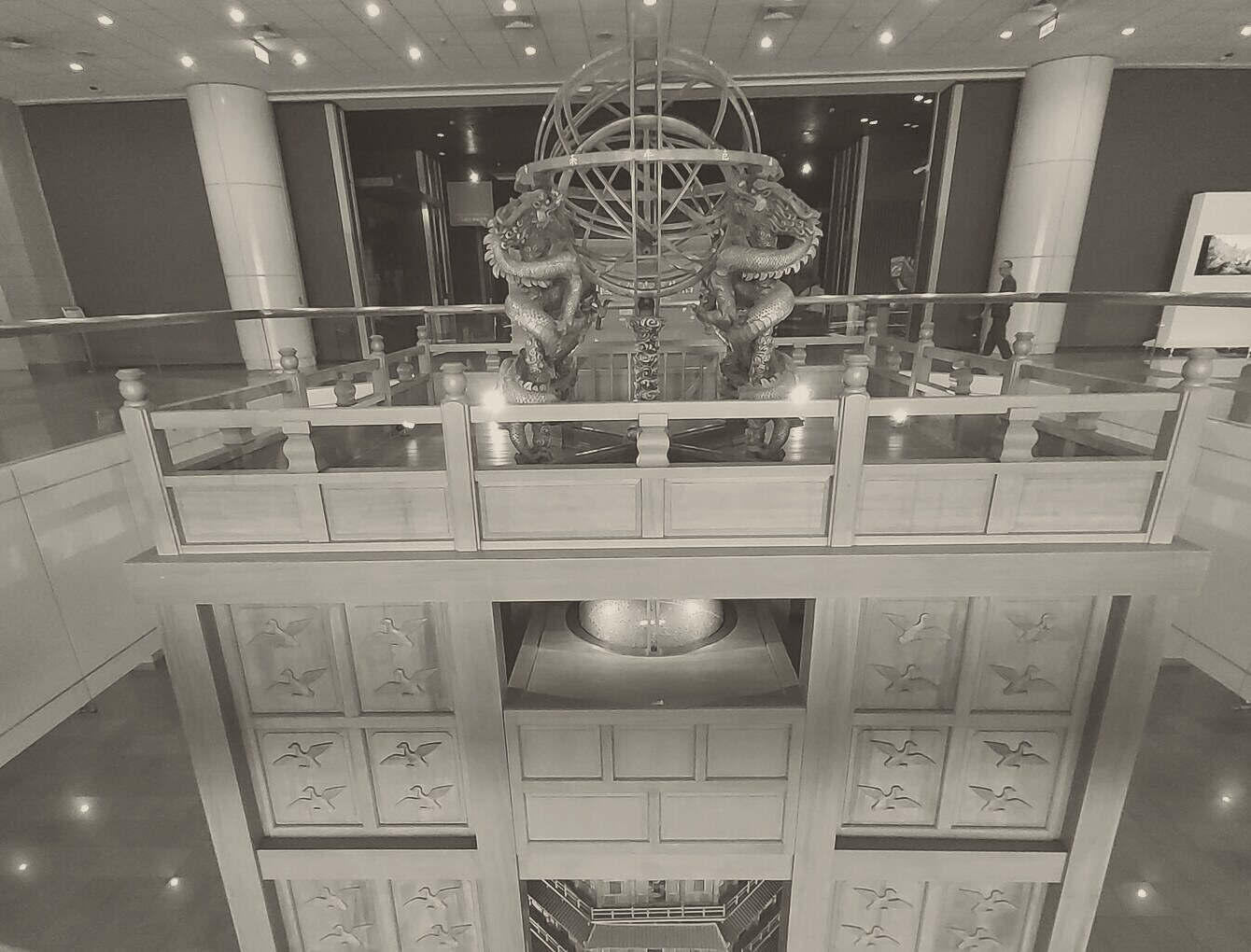 Looking down on the top of a tall wooden tower in a museum hall: an armillary sphere of interlocking metal rings stands on a stand of two coiled dragons inside a railed platform, and below it a lit, glowing globe sits in an open room in the tower's face.