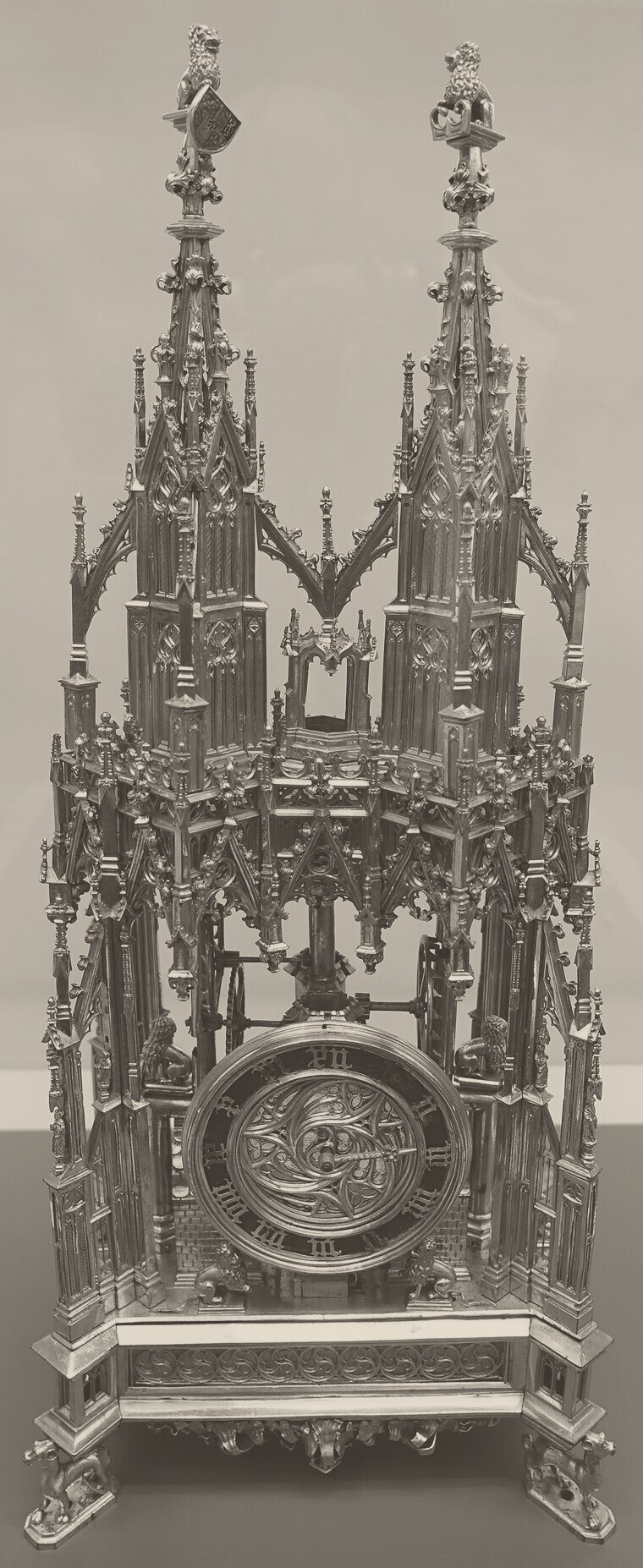 A tall gilded table clock shaped like a Gothic church, with two pointed spires topped by small lions, open tracery through which wheels can be seen, a round dial with Roman numerals and a single hand low down, and a box-like base standing on four small animal feet.