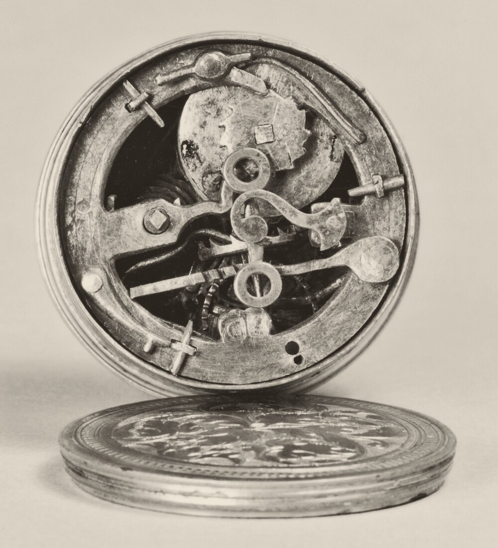 Black-and-white photograph of an early German drum watch standing open: a round, flat case with its lid lying in front of it, and inside the case a movement of plates, small wheels and a long curved spring arm.