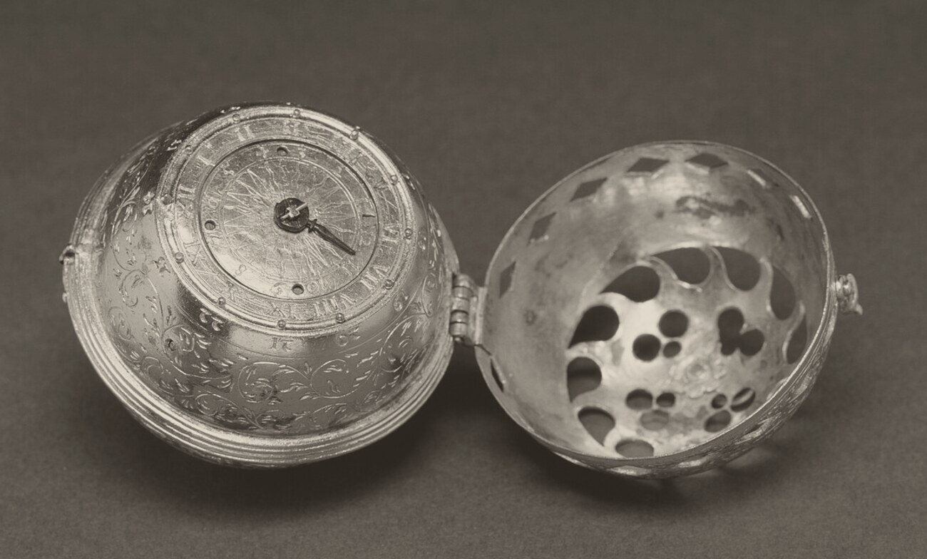 A small engraved metal ball opened on its hinge: the left half shows a round dial with Roman numerals and a single short hand; the right half, the lid, is pierced with round and diamond-shaped holes.