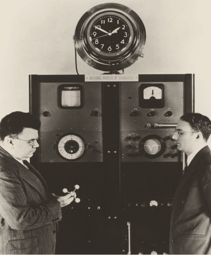 Black-and-white photograph of two men in suits either side of a tall cabinet of dials, meters and a small round oscilloscope screen, under a large wall clock and a plaque reading National Bureau of Standards. The man on the left holds a small model of a molecule.