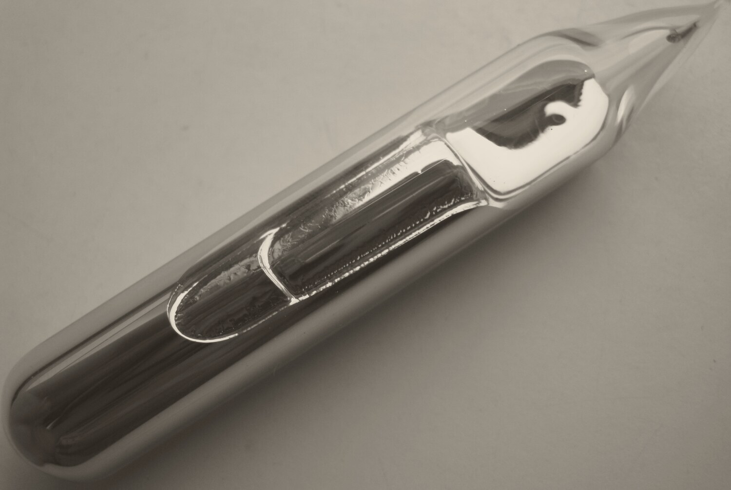 A sealed glass ampoule lying diagonally on a pale surface, its pointed tip at the top right, partly filled with a bright, mirror-like metal that has run along the inside of the glass.