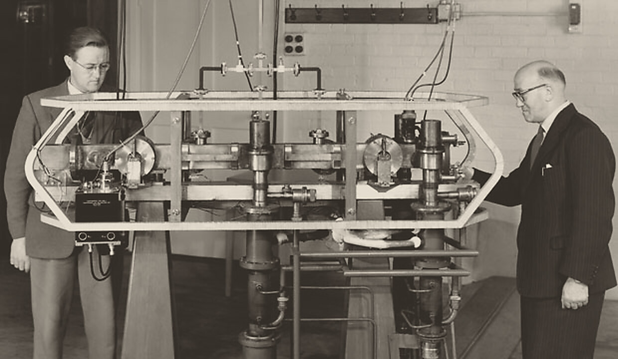 Black-and-white photograph of the first caesium clock at the National Physical Laboratory, 1955: a long horizontal tube of metal cylinders and valves inside an open six-sided frame, with Jack Parry standing at its left end and Louis Essen at its right, his hand on the frame.