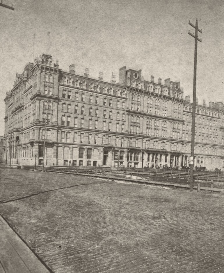 Old photograph of a long, six-storey stone hotel with ornate roof dormers and chimneys, seen across a wide cobbled street, with a telegraph pole in the foreground and carriages along the pavement.