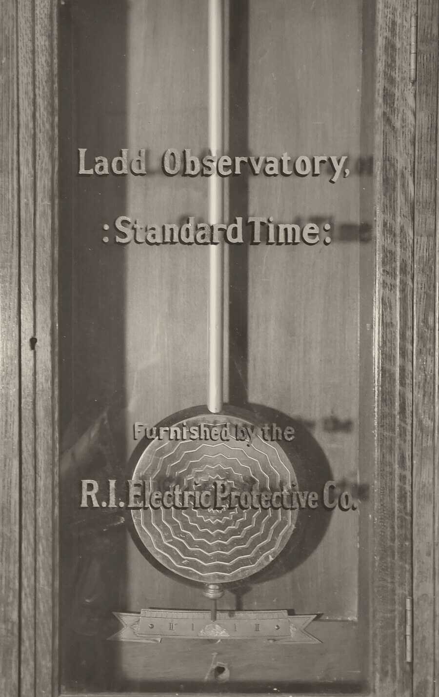 The glass door of a wooden clock case, lettered Ladd Observatory, Standard Time, Furnished by the R.I. Electric Protective Co., with a long pendulum rod and a round, patterned pendulum bob behind the glass.